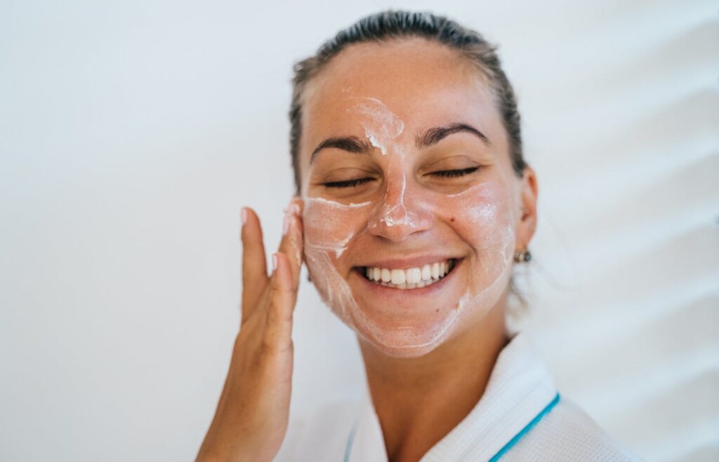 A smiling woman applying cream on face. - Products For Skin Barrier Repair